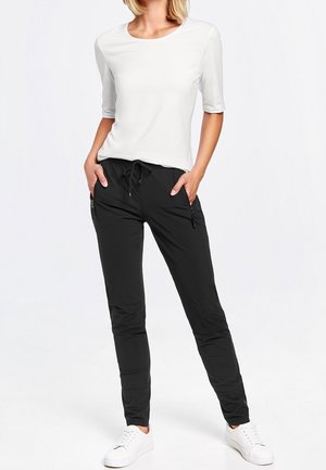 Femme portant un haut blanc à manches mi-longues, un pantalon noir à cordon avec poches zippées et des baskets blanches, debout les mains dans les poches.