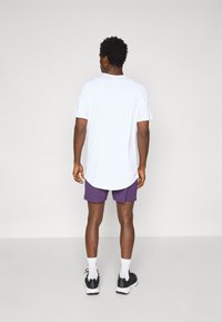 T-shirt blanc à manches courtes avec un ourlet arrondi, associé à un short violet. Le modèle porte des chaussures de sport noires et des chaussettes blanches, se tenant devant un fond uni.