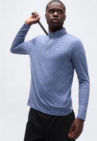 Άνδρας που κρατάει γκολφ κλαμπ πίσω από τον λαιμό, φοράει μπλε μπλουζάκι με μακριά μανίκια και φερμουάρ και μαύρο παντελόνι σε λευκό φόντο.