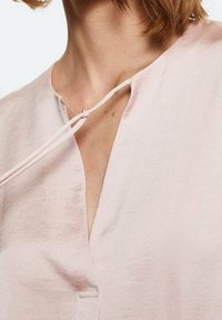 Blusa rosa chiaro con scollatura a V e chiusura con fiocco. Il tessuto appare liscio e leggermente lucido, con dettagli di design minimalisti.