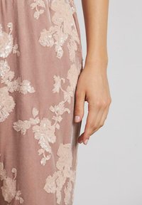Mano appoggiata accanto a un vestito di velluto rosa con ricami di paillettes floreali rosa chiaro su uno sfondo grigio semplice.