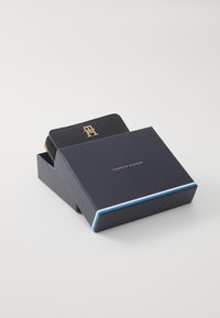 Juodas odinis piniginė, iš dalies matoma tamsiai mėlyname dėkle, su auksiniu logotipu ir užtrauktuko detalėmis; dėklas turi spalvotą juostelę krašte.