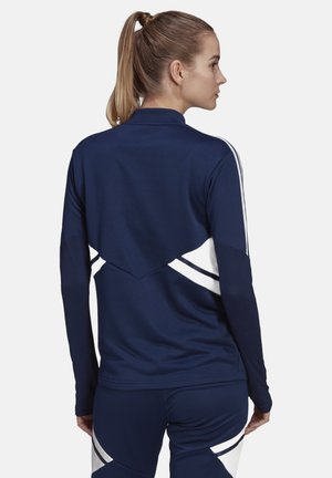 Marineblauwe sportjack met witte accenten, voorzien van een hoge kraag en een uniek ontwerp aan de achterkant. Soepele textuur, aansluitende mouwen en sportieve snit.