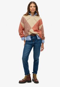 Suéter con un patrón de diamantes multicolores en beige, óxido y naranja. Combinado con una camisa de cuello azul claro y jeans azules. Botas marrones.