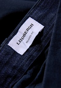 Marineblauwe stof met een visgraatpatroon; wit label met de tekst "LINDBERGH" en "RELAXED FIT." Zichtbare stiksels en textuurdetails.