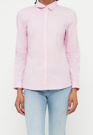 Femme portant une chemise rose clair à rayures verticales avec col et boutons, associée à un jean bleu clair, debout devant un fond blanc.