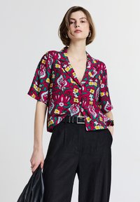 Camicia a maniche corte con bottoni in tessuto fucsia vibrante, caratterizzata da un motivo floreale e paisley. Abbinata a pantaloni neri a vita alta.