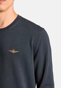 Maglione lavorato a maglia blu navy con scollatura rotonda, caratterizzato da una corona dorata e un emblema con ali sul lato sinistro del petto. Tessuto morbido, design aderente.