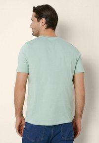 Tricou din bumbac de culoare verde mentă, cu mâneci scurte și decolteu rotund, cu o textură netedă și o croială relaxată, purtat cu pantaloni scurți din denim albastru.
