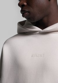 Lys beige hoodie med en afslappet pasform, der har en stor hætte og et diskret broderet logo i en matchende farve på brystet. Glat tekstur.