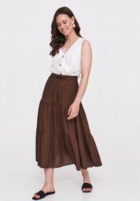 Biała bezrękawnik z guzikami, zestawiona z brązową, warstwową maxi spódnicą. Spódnica ma elastyczny pas i lekki połysk. Czarna sandały.
