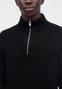 Personne portant un pull noir côtelé demi-zip, vue des épaules à la bouche, sur un fond uni clair.