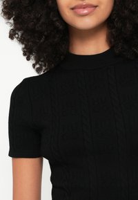 Μαύρο πλεκτό τοπ με κοντά μανίκια, διακοσμημένο με σχέδιο ριμπ και ανάγλυφα καλώδια, με λαιμόκοψη τύπου mock neck. Το υλικό φαίνεται μαλακό και με υφή.