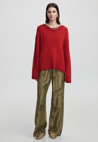 Roter Strickpullover mit strukturiertem Finish und einer Kettenakzent am Ausschnitt, kombiniert mit weiten olivgrünen Hosen mit floralem Muster.