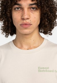 Camiseta de algodón beige con cuello redondo; presenta texto verde "Element Skateboard Co" en la parte frontal. Textura suave, ajuste casual.