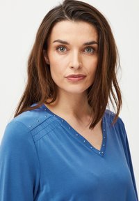 Kék, hosszú ujjú, V-nyakú felső, díszítő szegecsekkel a nyakvonal mentén és finom ráncolással a vállakon. Sima textúra.