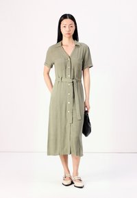 Robe chemise olive, boutonnée, à manches courtes, avec une ceinture à nouer et deux poches poitrine. Le modèle tient un sac noir et se tient sur un fond uni.