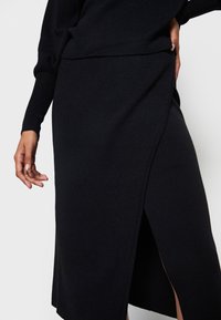 Jupe noire en maille ajustée avec une fente latérale. Le tissu est lisse, et le design présente une coupe épurée et moderne, mettant en valeur une silhouette élégante.