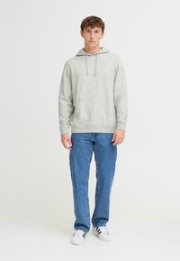 Sudadera gris con capucha y bolsillo frontal, combinada con jeans azules y zapatillas blancas. Diseño casual con un corte holgado.
