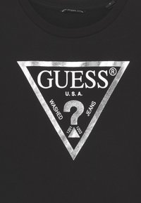 Černé bavlněné tričko s velkým stříbrným trojúhelníkovým logem, na kterém je napsáno "GUESS," "WASHED," "JEANS," a uprostřed je otazník.