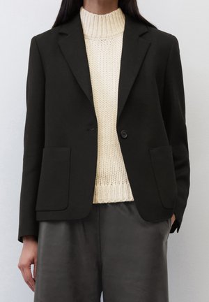 Kobieta ubrana w czarną marynarkę na kremowym swetrze z dzianiny i czarne skórzane spodnie, stojąca na tle jasnego, jednolitego tła.