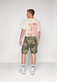 Beige graphic t-shirt with orange design on the back, paired with green camo cargo shorts. Model wears white sneakers and striped socks.