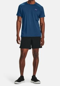 Blue athletic t-shirt with short sleeves, featuring a pink logo on the chest, paired with black shorts and black athletic shoes.