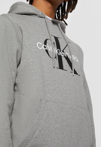 Szara bluza z kapturem, wykonana z miękkiego materiału, z przednią kieszenią i dużym czarnym logo "CK" z napisem "Calvin Klein" powyżej.