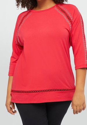 Vrouw die een rood top met driekwart mouwen met gaasuitsnijdingen en strassaccenten op de mouwen en zoom draagt, gecombineerd met een zwarte broek.