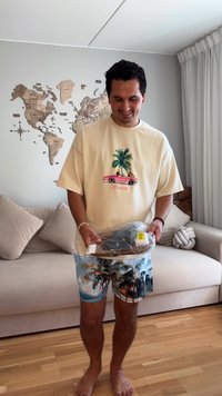 Beige T-Shirt mit einem pinken Cabrio, grünen Palmen und dem Wort "Miami". Bunte Shorts mit tropischem Muster, dabei verpackte Schuhe.