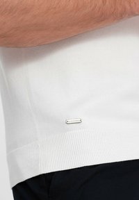 Pullover bianco lavorato a maglia con orli e polsini a coste, caratterizzato da un piccolo tag argento sulla parte laterale con testo inciso. Design e texture semplici.