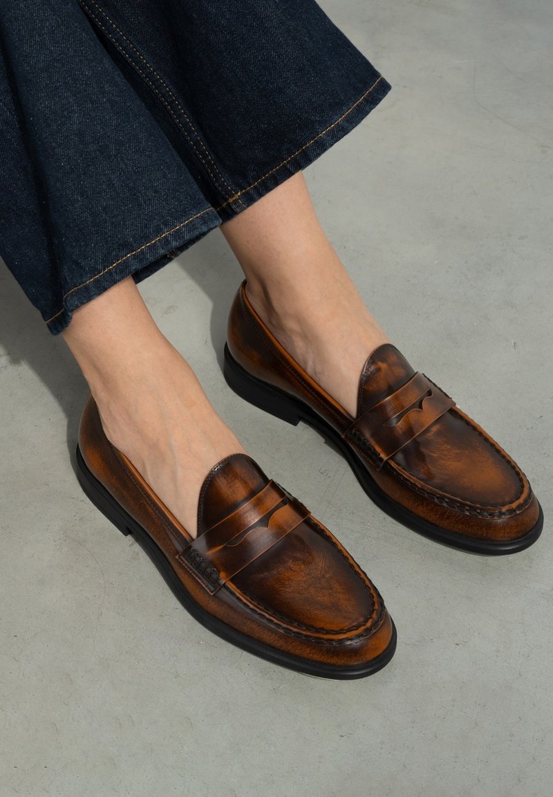 Brown leather loafers with a smooth texture, featuring a saddle strap detail and black rubber soles, worn with dark denim.