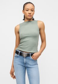 Νεαρή γυναίκα με πλεξούδες, φορώντας πράσινο ριμπ τοπ χωρίς μανίκια, ανοιχτόχρωμα μπλε τζιν και μαύρη ζώνη με μεγάλο διακοσμητικό αγκράφα σε σχήμα καρδιάς.