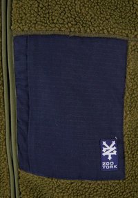 Fleecejacke mit einer olivgrünen Textur, mit einem marineblauen Panel und einem blauen Logo-Tag in der unteren Ecke. Sichtbares Reißverschlussdetail.