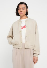 Μπεζ cropped μπουφάν από μαλακό ύφασμα με στρογγυλή λαιμόκοψη, χρυσά κουμπώματα και δύο τσέπες μπροστά. Τα μανίκια έχουν σχήμα μπαλονιού.