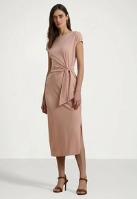 Žena v svetloružových šatách po kolená so šnúrkou na boku a hnedými sandálmi na vysokom opätku, stojaca na neutrálnom pozadí.