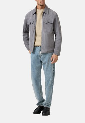 Man wearing light gray suede jacket, beige knit sweater, light blue jeans, and black loafers, standing with hands relaxed.