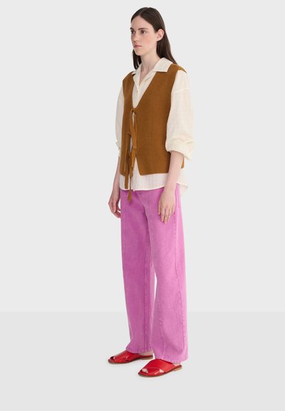 Femme portant un chemisier crème à manches bouffantes, un gilet tricoté marron noué devant, un pantalon large rose et des sandales rouges à bout ouvert.