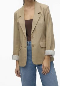 Beige blazer with notched lapels, two buttons, and front pockets. Cuffed sleeves with a white striped pattern. Paired with light blue jeans.