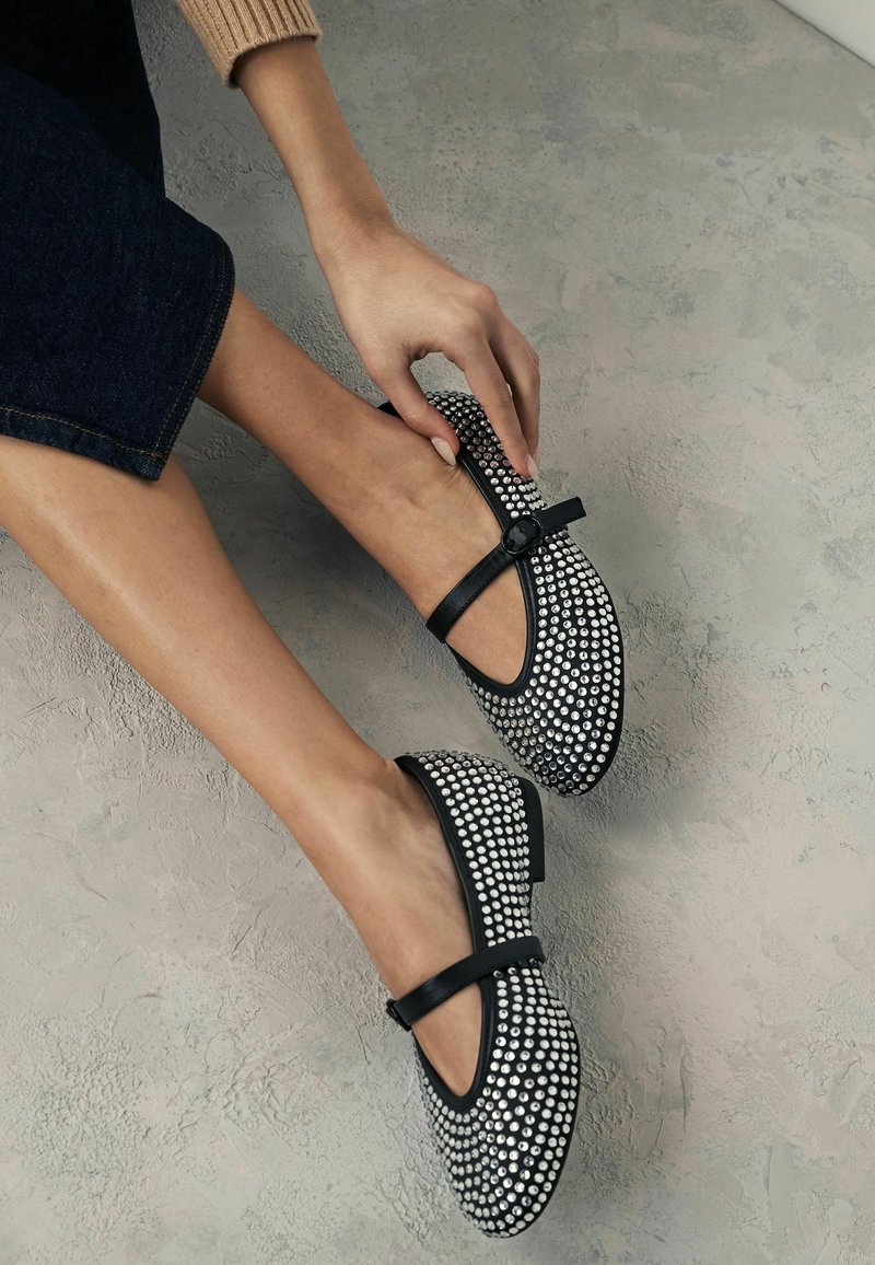 Ballerines noir et blanc avec un bout rond, des embellissements décoratifs à clous, et une sangle noire sur le cou-de-pied.