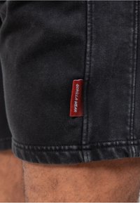 Zwarte denim short met zichtbare steken, met een rood label dat "GORILLA WEAR" leest. De stof heeft een gestructureerde uitstraling.