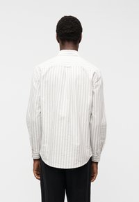 Άνδρας που φοράει μακρυμάνικο, ανοιχτόχρωμο πουκάμισο με ρίγες, στέκεται γυρισμένος με την πλάτη προς μια απλή λευκή επιφάνεια.