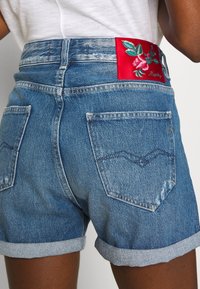 Světle modré džínové šortky s ohrnutým lemem. Na zadním pasu mají červenou vyšívanou nášivku s květinovým motivem a dvě zadní kapsy.