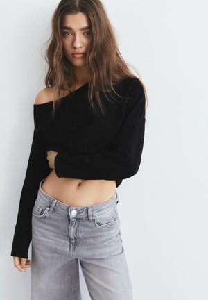 Ung kvinde med langt brunt hår iført en sort off-shoulder cropped sweater og grå højtaljede jeans mod en ensartet lys baggrund.