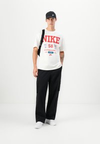 White cotton t-shirt with red graphic print, black cargo pants, and white sneakers. Black cap and crossbody bag complete the outfit.