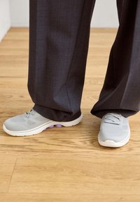 Chaussures de sport gris clair avec texture en maille et design à lacets, dotées d'une semelle blanche avec des accents violets. Portées avec un pantalon large et foncé.