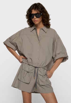 Mujer con camisa beige de gran tamaño abotonada con mangas remangadas, pantalones cortos a juego con grandes bolsillos delanteros y gafas de sol negras.
