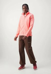 Naine seisab, seljas korallpunane tõmblukk-tuuletõke, pruunid püksid ja punased matkajalanõud, lameda heleda tausta ees.