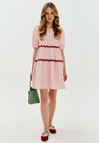 Różowa sukienka z krótkim rękawem z czerwonymi falbanami w talii, zwężany krój, w parze z zieloną torebką i czerwonymi balerinkami. Gładka faktura.