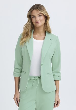 Kvinde med langt blondt hår iført en lysegrøn blazer over en hvid top og matchende lysegrønne bukser med snøre i taljen.